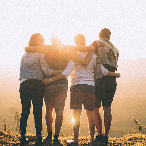 Group of four people standing together with arms around each other, facing a sunset